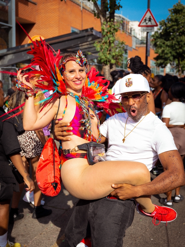 A Man In A White Cap Lifting A Woman Wearing A Vibrant, Feathered Carnival Costume On A Sunny Street.